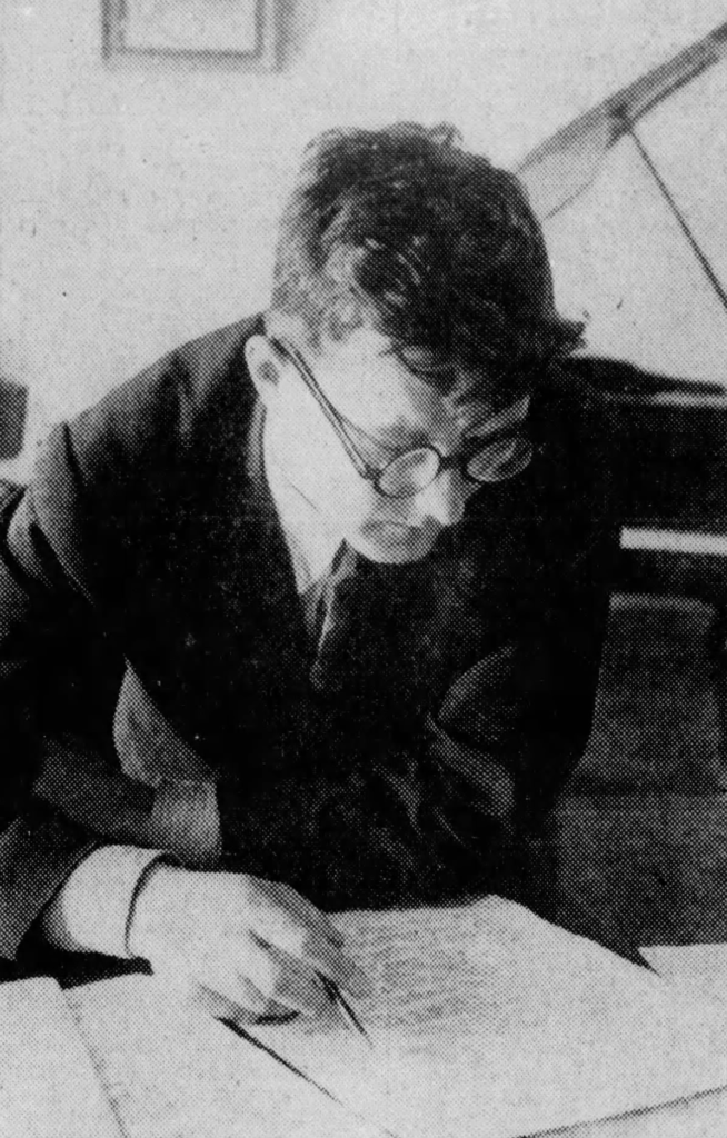 Schostakowitsch, mit einem Stift über eine Partitur gelehnt, hinter ihm ein Klavier bzw. Flügel, Anfang der 1940er Jahre