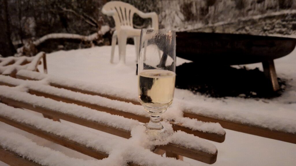Ein halbvolles Sektglas auf einem beschneiten Schlitten vor einer Feuerschale. Das Feuer ist für den Moment erloschen. 